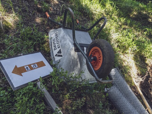 Am grünen Hang liegen Schubkarre, ein Wegschild sowie einen Drahtzaun als Arbeitsgeräte bereit. | © Davos Klosters Mountains