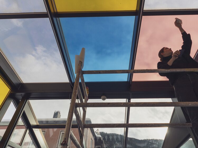 Eine Mitarbeiterin der Agentur Pfeffermint arbeitet an der Umgestaltung des Wintergartens und beklebt die Scheiben mit farbigen Folien.  | © Davos Klosters Mountains