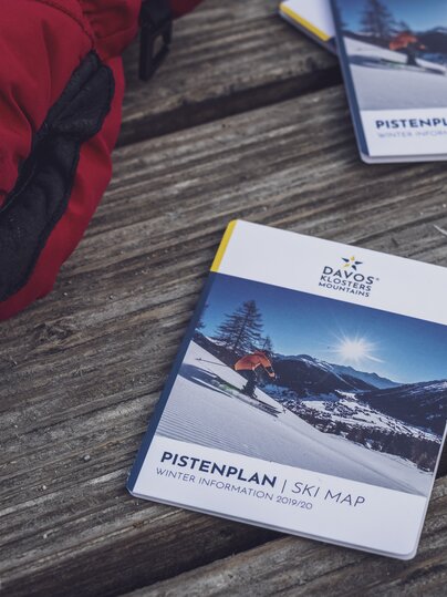 Pistenpläne und eine roter Handschuh liegen auf dunklem Holztisch. | © Davos Klosters Mountains