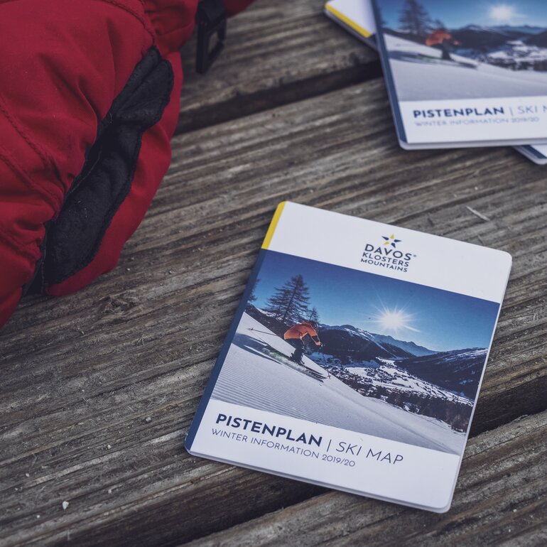 Ski maps and a red glove lie on a dark wooden table. | © Davos Klosters Mountains