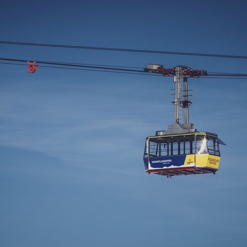 Vor blauem Himmel schwebt die blau/gelbe Gondel der Gipfelbahn zum Weissfluhgipfel auf Parsenn.   | © Davos Klosters Mountains