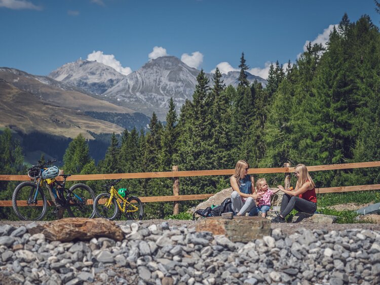 Zwei Frauen und ein Mädchen sitzen am Ufer eines Bergsees, daneben steht ein grosses und ein kleines Bike. | © Davos Klosters Mountains