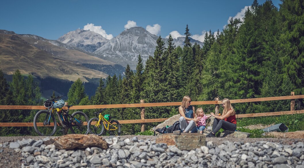 Familie am Bergsee mit Mountainbikes | © Davos Klosters Mountains Zwei Frauen und ein Mädchen sitzen am Ufer eines Bergsees, daneben steht ein grosses und ein kleines Bike. | © Davos Klosters Mountains