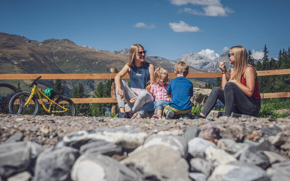 Zwei Frauen und zwei Kinder sitzen am See auf dem Rinerhorn und stärken sich mit einem Snack. | © Davos Klosters Mountains