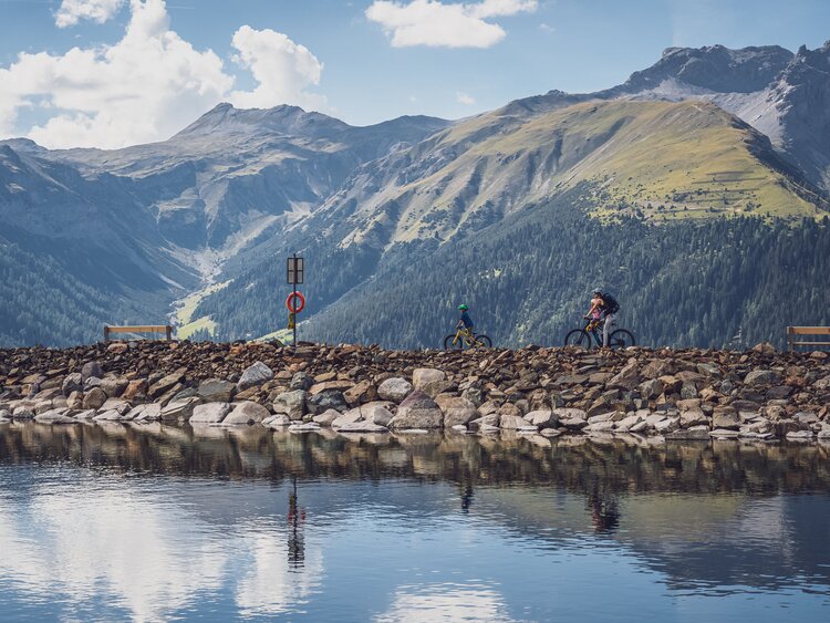 Eine Frau fährt mit ihren beiden Kindern mit dem Mountainbike am Seeufer entlang. | © Davos Klosters Mountains