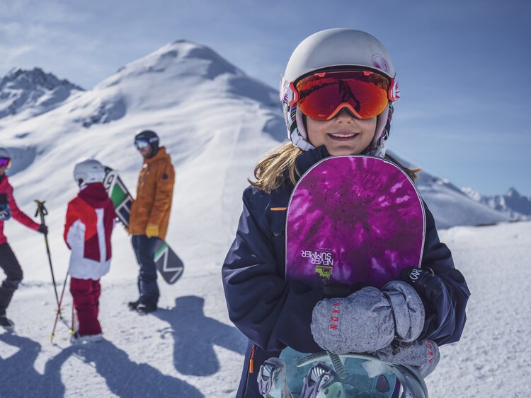Junges Mädchen mit Never Summer Snowboard in der Hand und ihrer Famile im Hintergrund. | © Davos Klosters Mountains