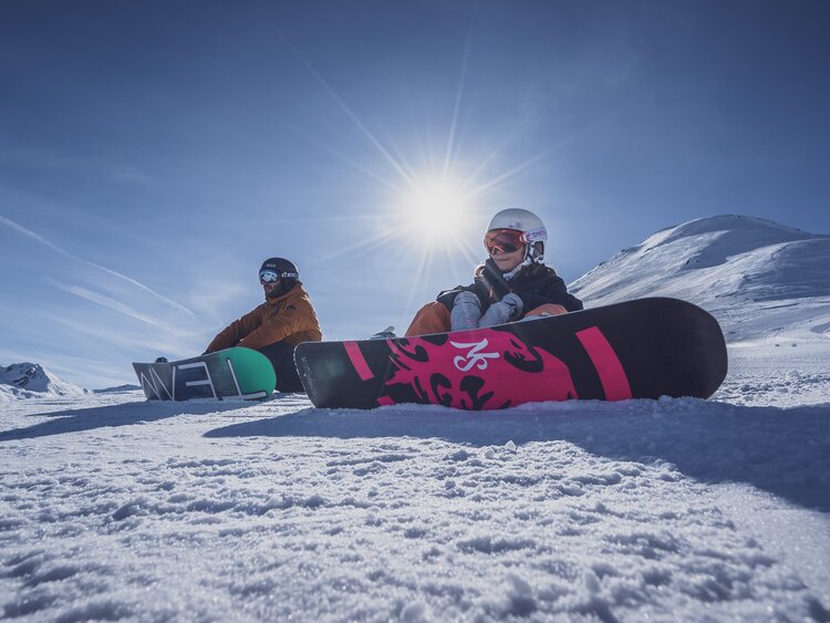 Vater und Tochter sitzen mit Snowboard angeschnallt auf der Piste bei Sonnenschein. | © Davos Klosters Mountains