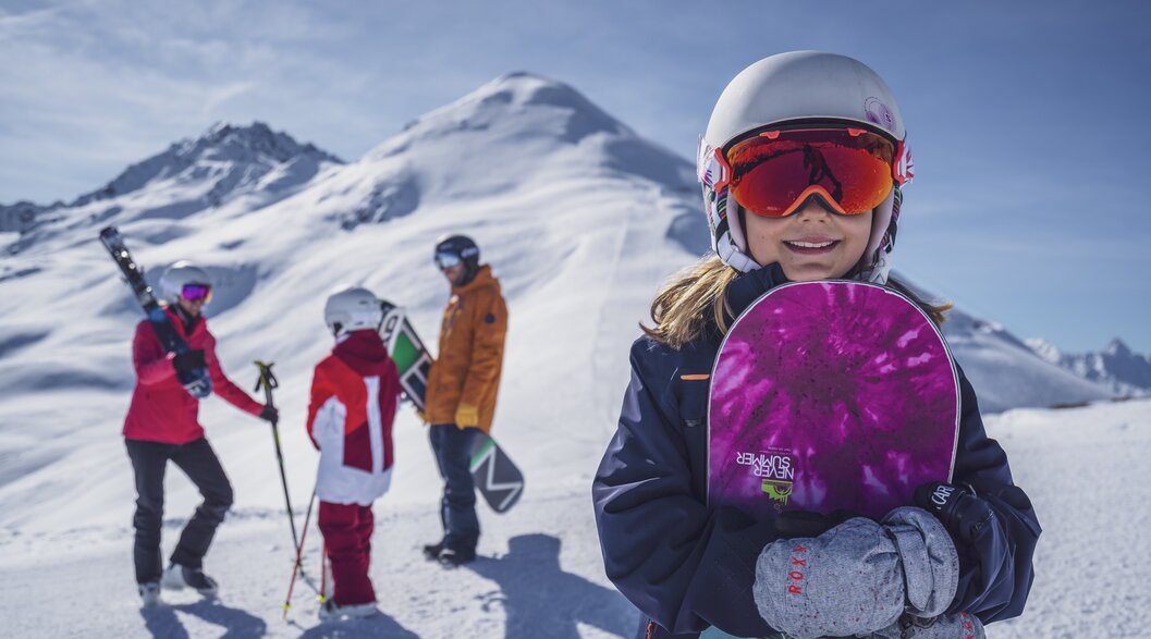 Snowboarder Mädchen | © Davos Klosters Mountains Junges Mädchen mit Never Summer Snowboard in der Hand und ihrer Famile im Hintergrund. | © Davos Klosters Mountains