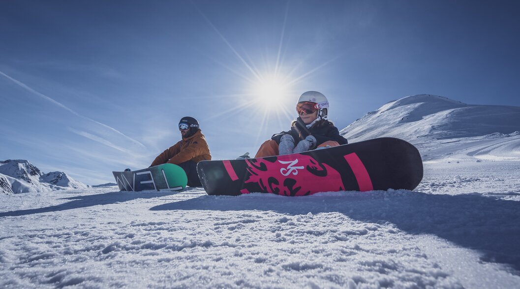 Vater-Tochter Snowboard Tag | © Davos Klosters Mountains Vater und Tochter sitzen mit Snowboard angeschnallt auf der Piste bei Sonnenschein. | © Davos Klosters Mountains