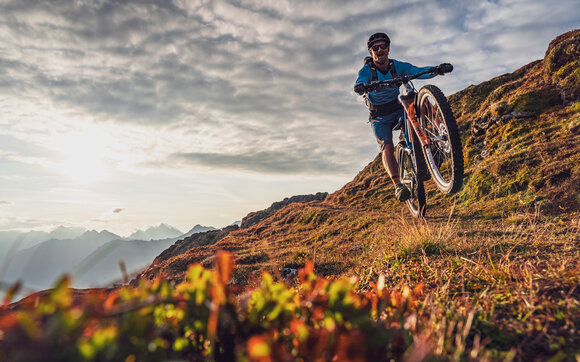 Biker fährt durch eine herbstliche Landschaft im goldenen Licht und hebt sein Vorderrad an. | © Davos Klosters Mountains