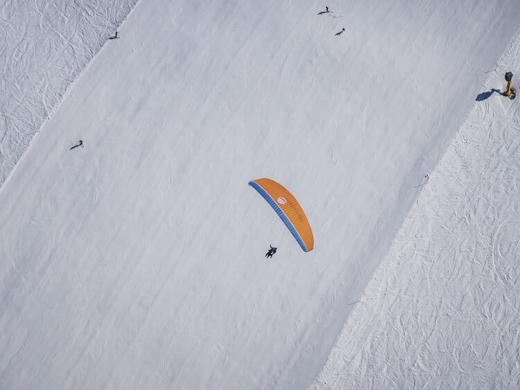 Ein oranger Gleitschirm überfliegt eine Skipiste, die in gerader Linie unter ihm liegt.  | © Davos Klosters Mountains