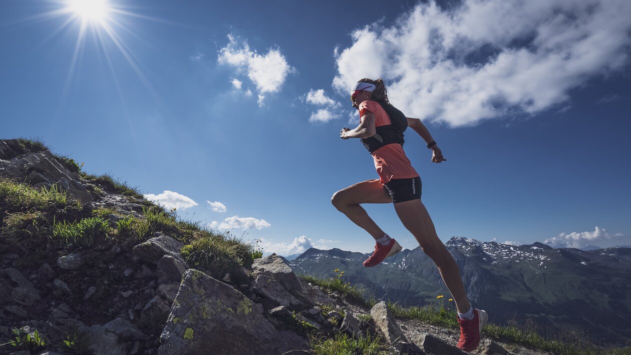 Mit dynamischer Körperspannung rennt die Trailrunnerin den Berg hoch, mit der Sonne am Himmel. | © Davos Klosters Mountains