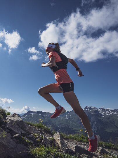 Mit dynamischer Körperspannung rennt die Trailrunnerin den Berg hoch, mit der Sonne am Himmel. | © Davos Klosters Mountains