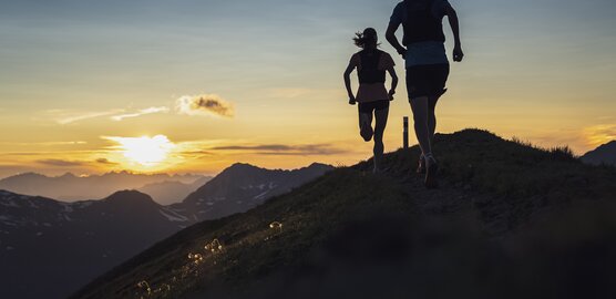Zwei Trailrunner rennen auf dem Grat des Jakobshorn in Richtung Sonnenuntergang mit orange-farbenem Himmel.