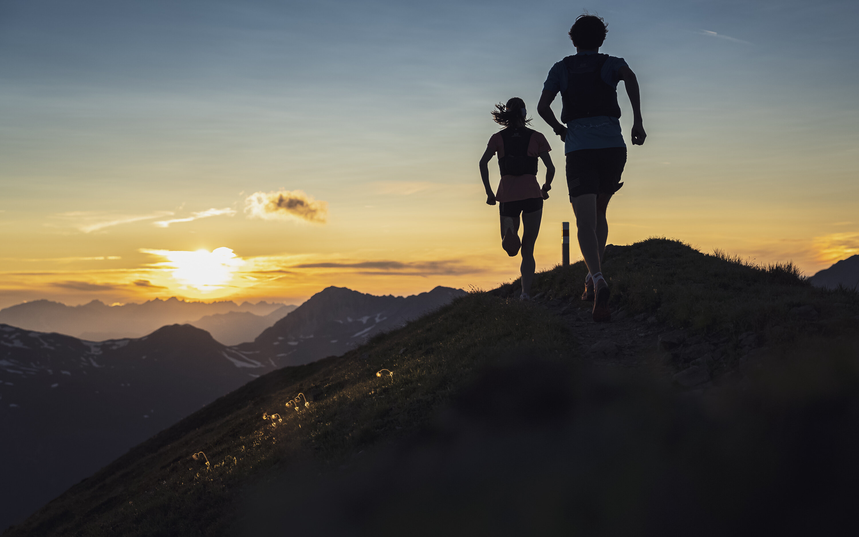 Zwei Trailrunner rennen auf dem Grat des Jakobshorn in Richtung Sonnenuntergang mit orange-farbenem Himmel. | © Davos Klosters Mountains