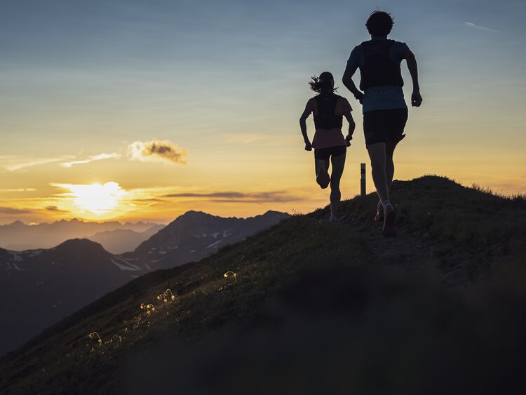Zwei Trailrunner rennen auf dem Grat des Jakobshorn in Richtung Sonnenuntergang mit orange-farbenem Himmel. | © Davos Klosters Mountains