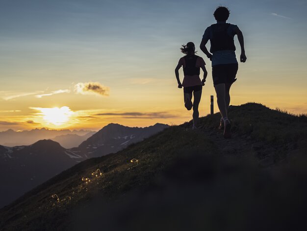 Zwei Trailrunner rennen auf dem Grat des Jakobshorn in Richtung Sonnenuntergang mit orange-farbenem Himmel. | © Davos Klosters Mountains
