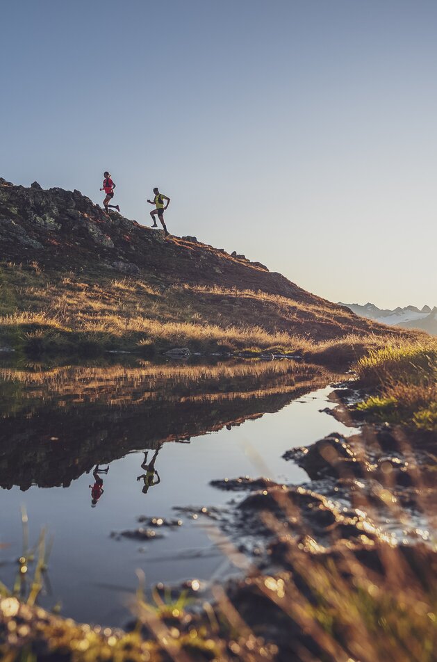 Zwei Trailrunner sind bei Sonnenaufgang laufend auf Gotschna unterwegs und werden im See gespiegelt. | © Davos Klosters Mountains