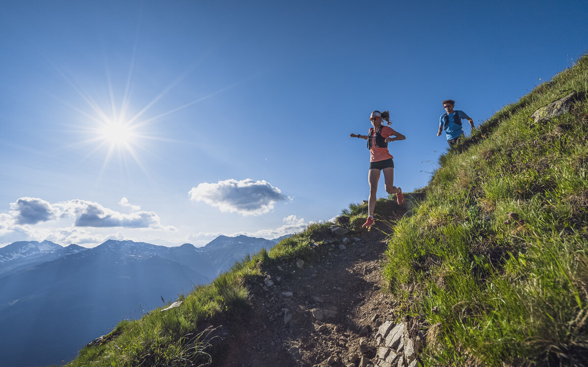 Eine Trailrunnerin springt bei sonnigem Wetter den Weg vom Brämabüel zum Berg Jakobshorn hinab, es folgt Ihr ein Trail Runnern in blauem T-Shirt.  | © Davos Klosters Mountains