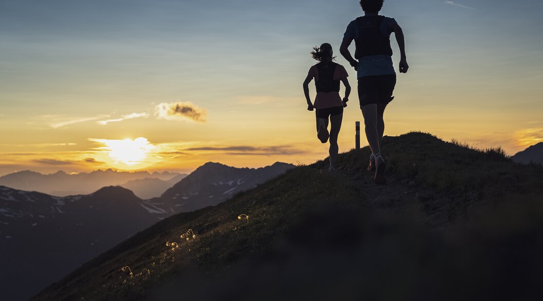 Trailrunning Paar Sonnenuntergang Jakobshorn | © Davos Klosters Mountains Zwei Trailrunner rennen auf dem Grat des Jakobshorn in Richtung Sonnenuntergang mit orange-farbenem Himmel. | © Davos Klosters Mountains