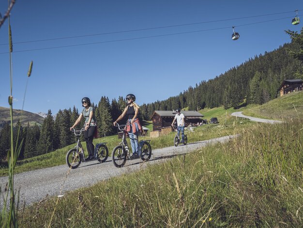 Eine Gruppe von Freunden fährt mit den Trottinette unter der Gondelbahn Rinerhorn hindurch, vorbei an einer  grünen Wiese in Richtung Tal.  | © Davos Klosters Mountains