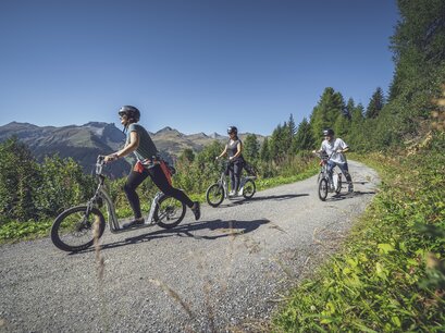 Zwei Trottinettfahrer geben mit den Beinen Schwung um Tempo zu gewinnen, die Dritte steht auf dem Gefährt und geniesst die Abfahrt auf dem Trottinettweg auf dem Rinerhorn.  | © Davos Klosters Mountains