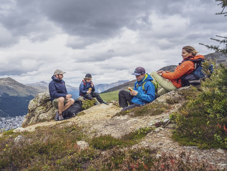 Eine vierköpfige Familie legt auf Ihrer Wanderung auf dem Rinerhorn eine Pause ein und hat sich für die Stärkung einen Platz mit Blick nach Davos ausgesucht.  | © Davos Klosters Mountains