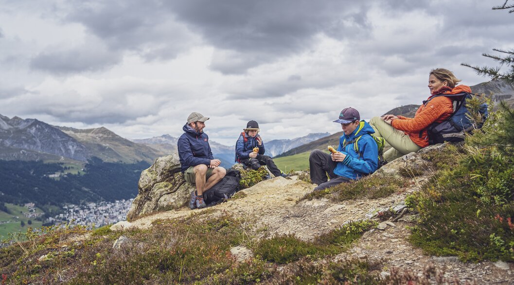 A family of four is taking a break on their hike on the Rinerhorn and has chosen a spot with a view of Davos for their rest.  | © Davos Klosters Mountains