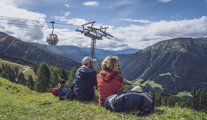 A family sits on the mountain meadow on the Rinerhorn and enjoys nature, behind them a gondola of the Rinerhorn cable car floats towards the mountain.  | © Davos Klosters Mountains