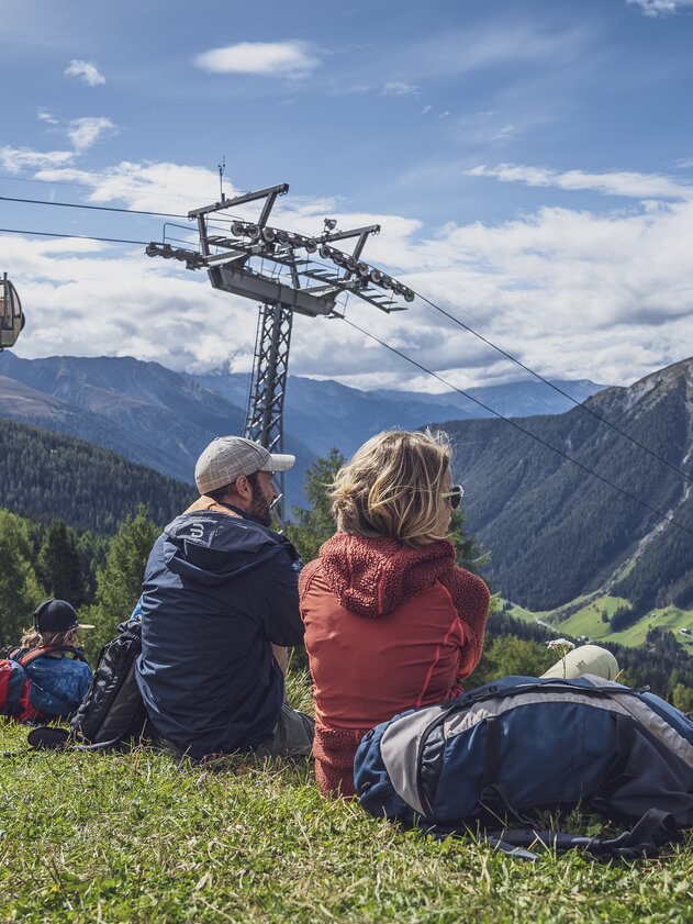 Wanderung Familie Rinerhorn Gondel | © Davos Klosters Mountains Eine Familie sitzt auf der Bergwiese auf dem Rinerhorn und geniesst die Natur, dahinter schwebt eine Gondel der Rinerhornbahn in Richtung Berg. | © Davos Klosters Mountains