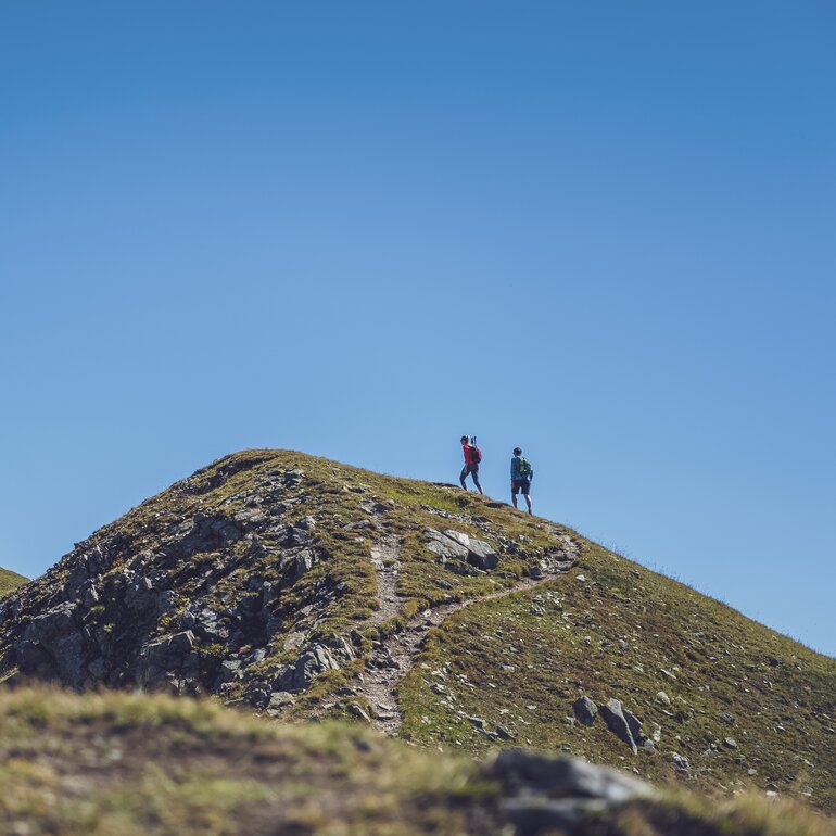 In der Ferne wandert ein Paar auf dem Grat des Jakobshorns in Richtung des Jatzhorns und geniesst den wolkenlosen Himmel.  | © Davos Klosters Mountains