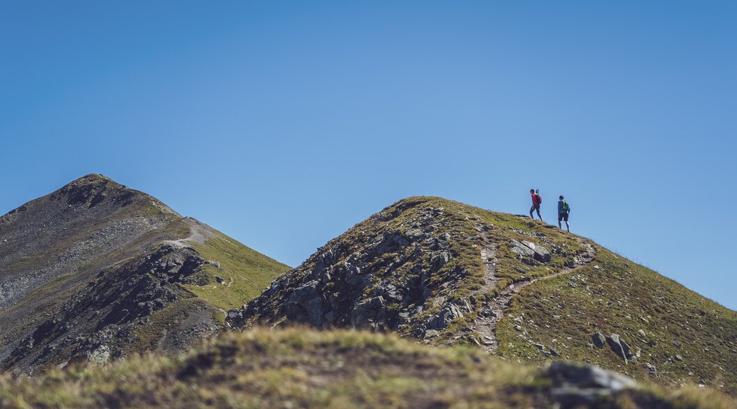 Wanderung Paar Jatzhorn | © Davos Klosters Mountains In der Ferne wandert ein Paar auf dem Grat des Jakobshorns in Richtung des Jatzhorns und geniesst den wolkenlosen Himmel. | © Davos Klosters Mountains