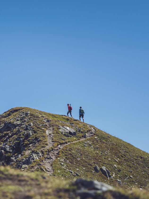 In der Ferne wandert ein Paar auf dem Grat des Jakobshorns in Richtung des Jatzhorns und geniesst den wolkenlosen Himmel.  | © Davos Klosters Mountains