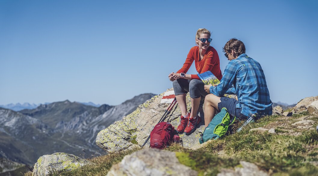 A couple sits on a stone on the summit, during the rest the man studies the hiking map and the woman enjoys the sun.  | © Davos Klosters Mountains