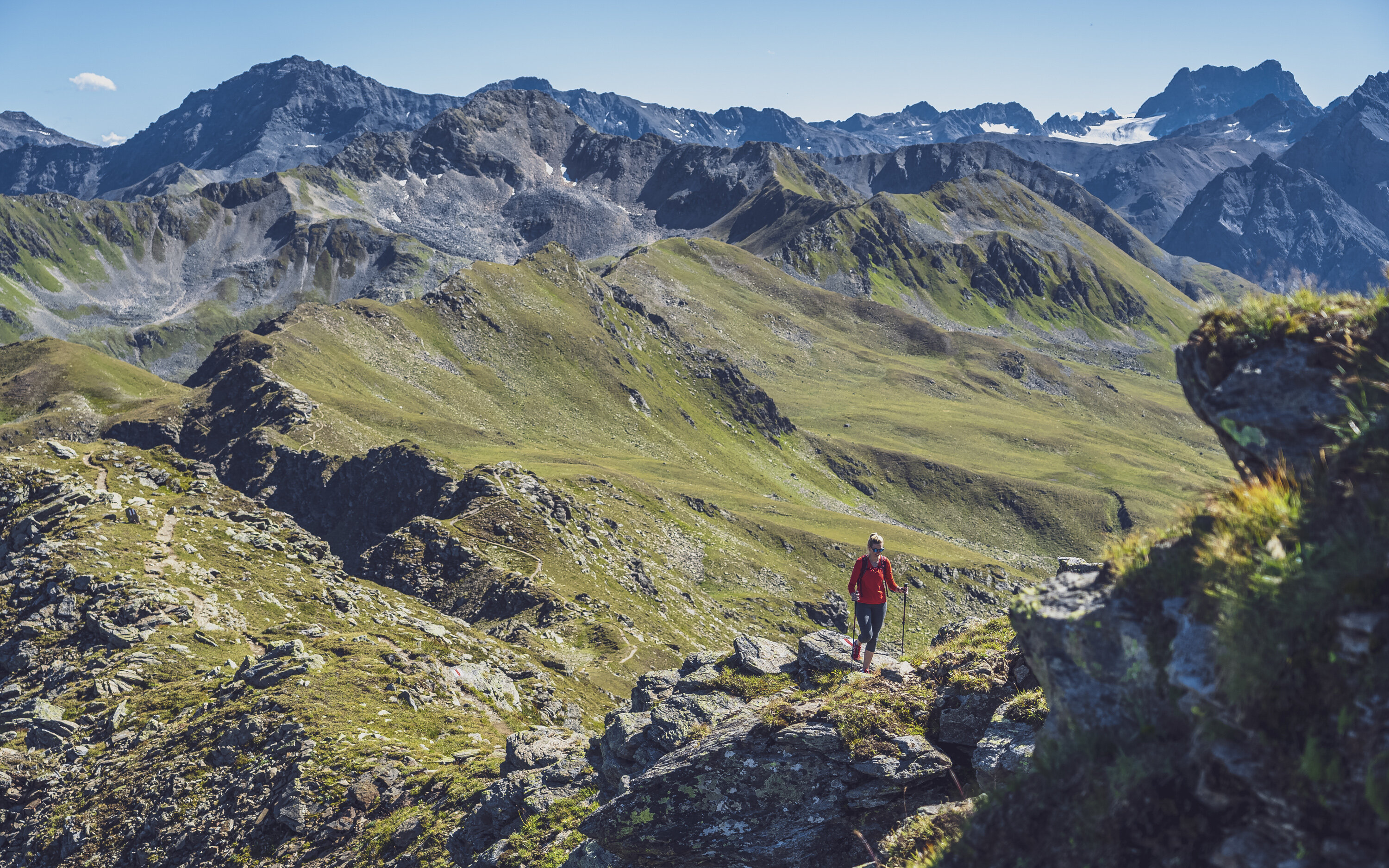 Eine in rot gekleidete Wanderin steigt mit ihren Stöcken den steinigen Grat hinauf, hinter ihr öffnet sich der Blick in das Sertig Tal.  | © Davos Klosters Mountains