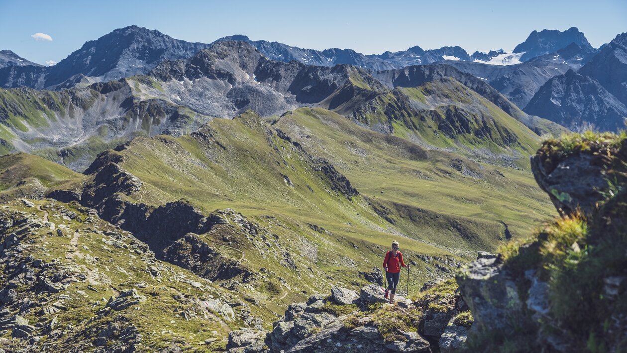 Eine in rot gekleidete Wanderin steigt mit ihren Stöcken den steinigen Grat hinauf, hinter ihr öffnet sich der Blick in das Sertig Tal.  | © Davos Klosters Mountains