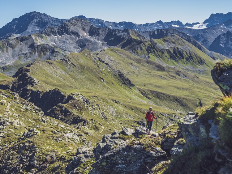 Eine in rot gekleidete Wanderin steigt mit ihren Stöcken den steinigen Grat hinauf, hinter ihr öffnet sich der Blick in das Sertig Tal.  | © Davos Klosters Mountains