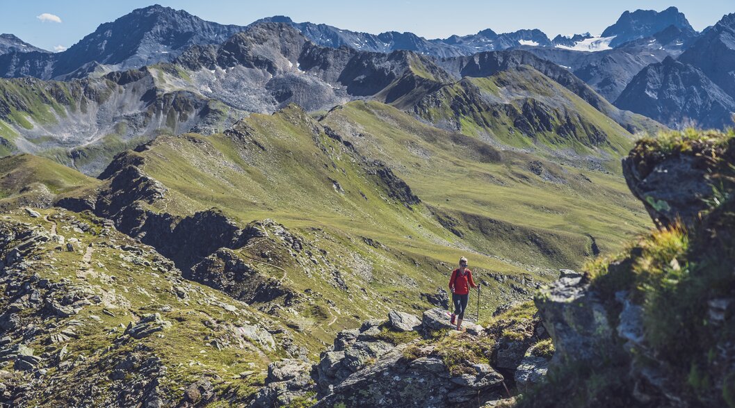 A female hiker dressed in red climbs up the stony ridge with her walking sticks, behind her the valley of Sertig is in view.  | © Davos Klosters Mountains