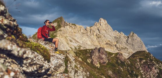 Ein Wanderer sitzt auf einem Felsen und blickt mit dem Fernglas in die Ferne.