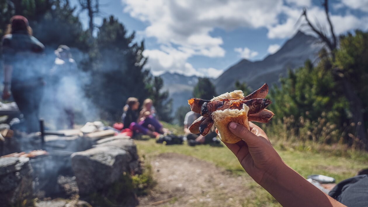 Ein Kind hält sein Brötchen mit der eingeklemmten Cervelat in die Höhe, dahinter die Grillstelle. | © Davos Klosters Mountains