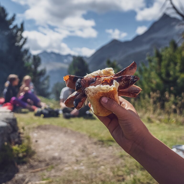Ein Kind hält sein Brötchen mit der eingeklemmten Cervelat in die Höhe, dahinter die Grillstelle. | © Davos Klosters Mountains