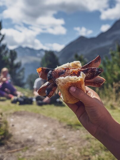 Ein Kind hält sein Brötchen mit der eingeklemmten Cervelat in die Höhe, dahinter die Grillstelle. | © Davos Klosters Mountains