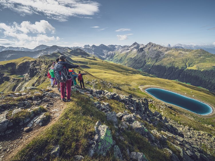 Eine Gruppe von Wanderer blickt vom Grat hinab auf den tiefblauen Stadlersee. | © Davos Klosters Mountains