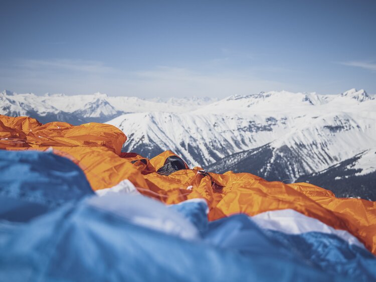 Nahaufnahme eines blau orangen Gleitschirmes der vor verschneiten Bergen am Boden ausgelegt ist.  | © Davos Klosters Mountains