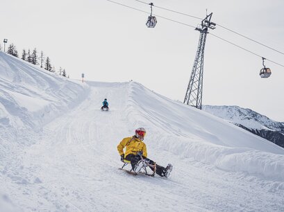Zwei Freundinnen schlitteln auf der Schlittelstrecke Rinerhorn um eine der 24 Kurve mit der Rinerhornbahn im Hintergund. | © Davos Klosters Mountains