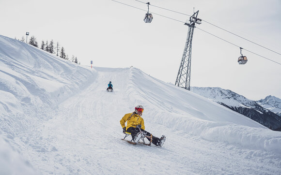 Two friends sledding on the Rinerhorn toboggan run around one of the 24 curves with the Rinerhorn cable car in the background. | © Davos Klosters Mountains