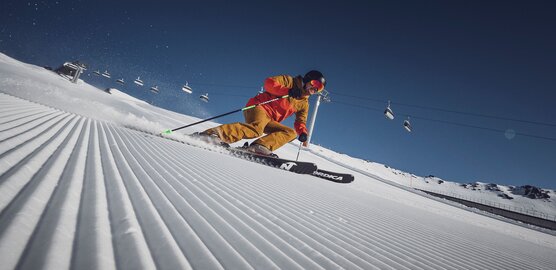 Ein Skifahrer am carven vor der Liftanlage auf einer perfekt präparierten Piste in der Sonne
