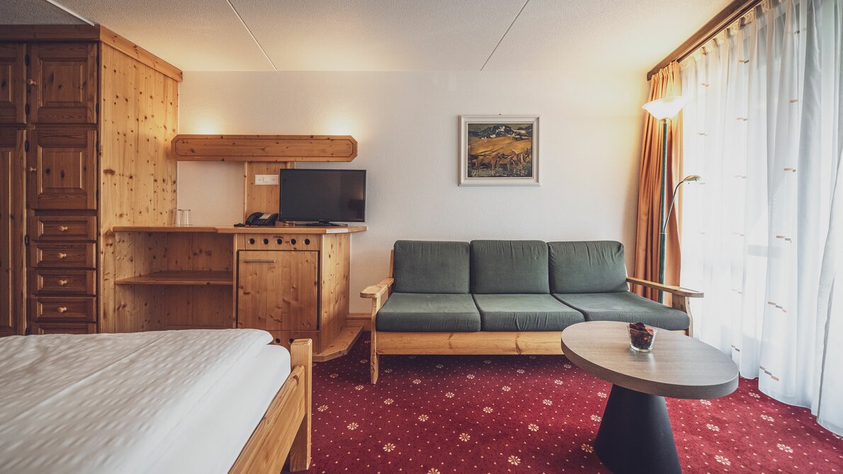 Double room with red carpet, window and curtains, sofa with small table, pine wood interior | © Davos Klosters Mountains Double room with red carpet, window and curtains, sofa with small table, pine wood interior | © Davos Klosters Mountains