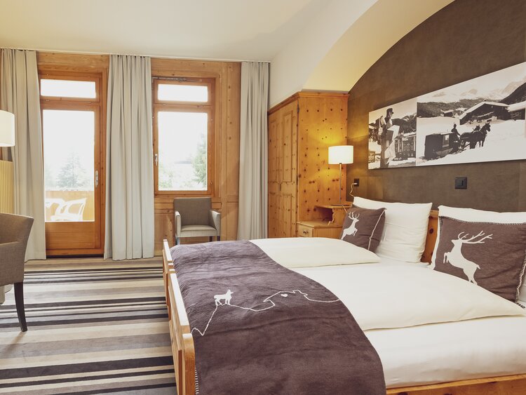 Doppelbettzimmer mit Bettwäsche im Hirschstyle, Fenster mit Stuhl und Holzeinrichtung | © Davos Klosters Mountains 