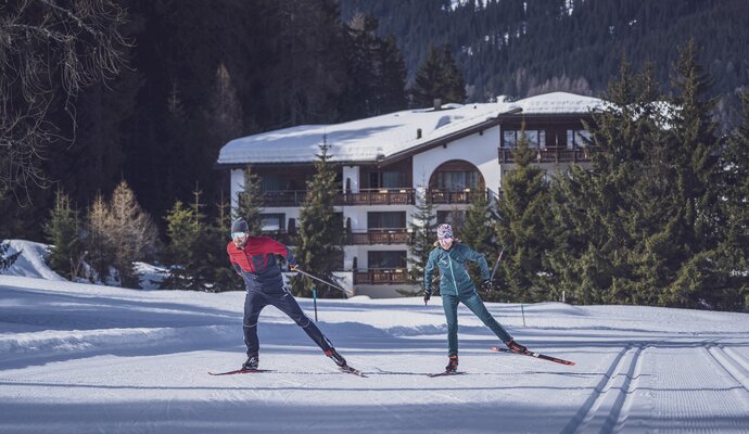 Two cross-country skiers on the trail with a hotel in the forest in the background | © Davos Klosters Mountains